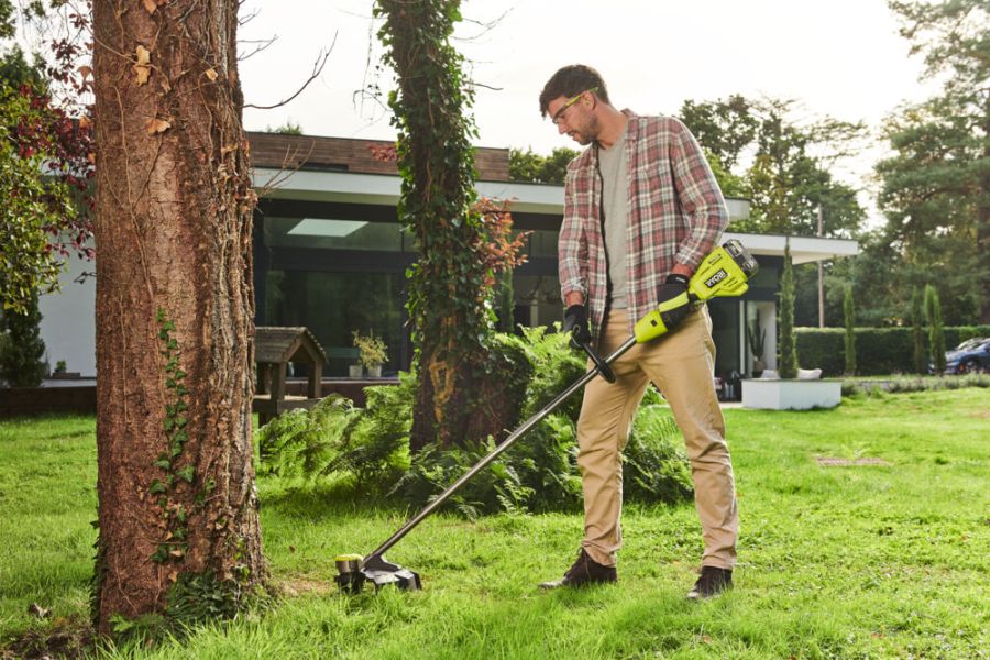 Mężczyzna używa podkaszarki RYOBI Podkaszarka 38cm bezszczotkowa WHISPER 18V ONE+ HP do cięcia trawy przy drzewie